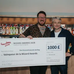 Two men at a bar holding a giant Gabriel Boudier Wizard Awards 2025 cheque for 1000 pounds, liqueur bottles on the shelf behind.