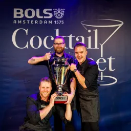 Three smiling bartenders in black aprons hold a large silver Bols Around the World trophy in front of a Bols Cocktail banner.